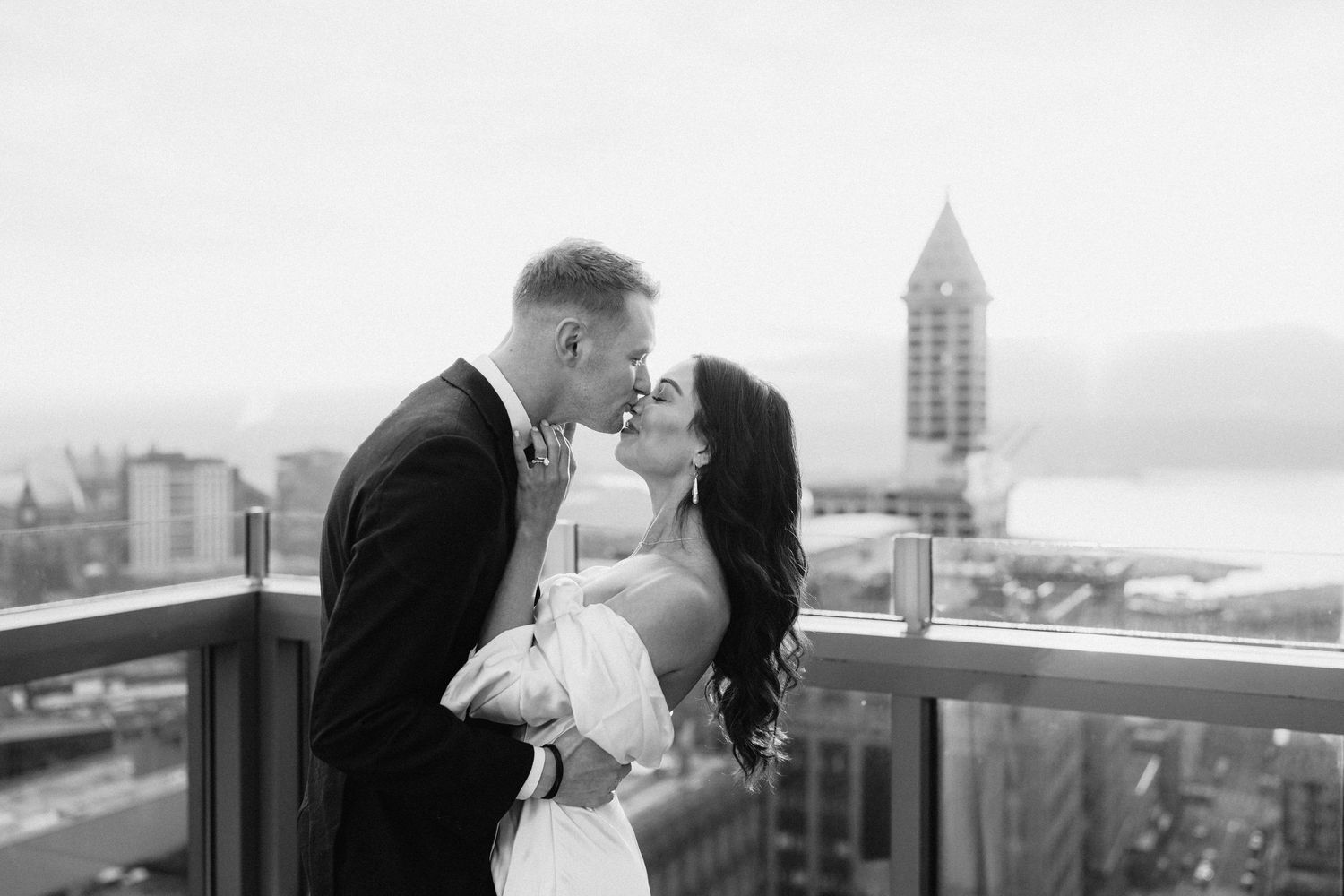 Black and white cityscape photo with couple sharing romantic moment on rooftop overlooking Seattle's Smith Tower and waterfront.