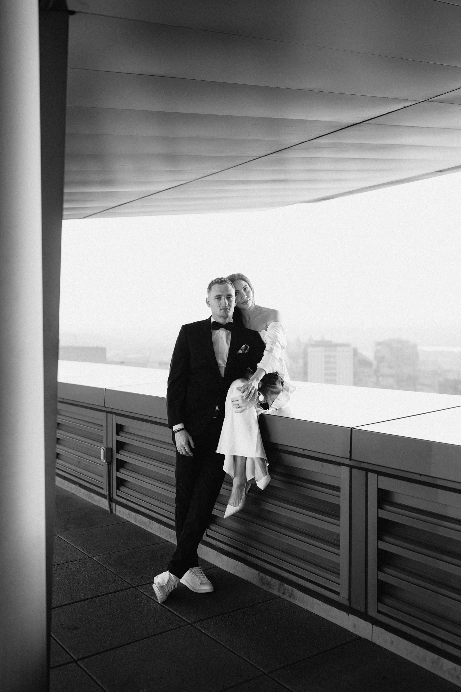 A black and white photo of a couple in formal attire standing together on a balcony with a cityscape view.