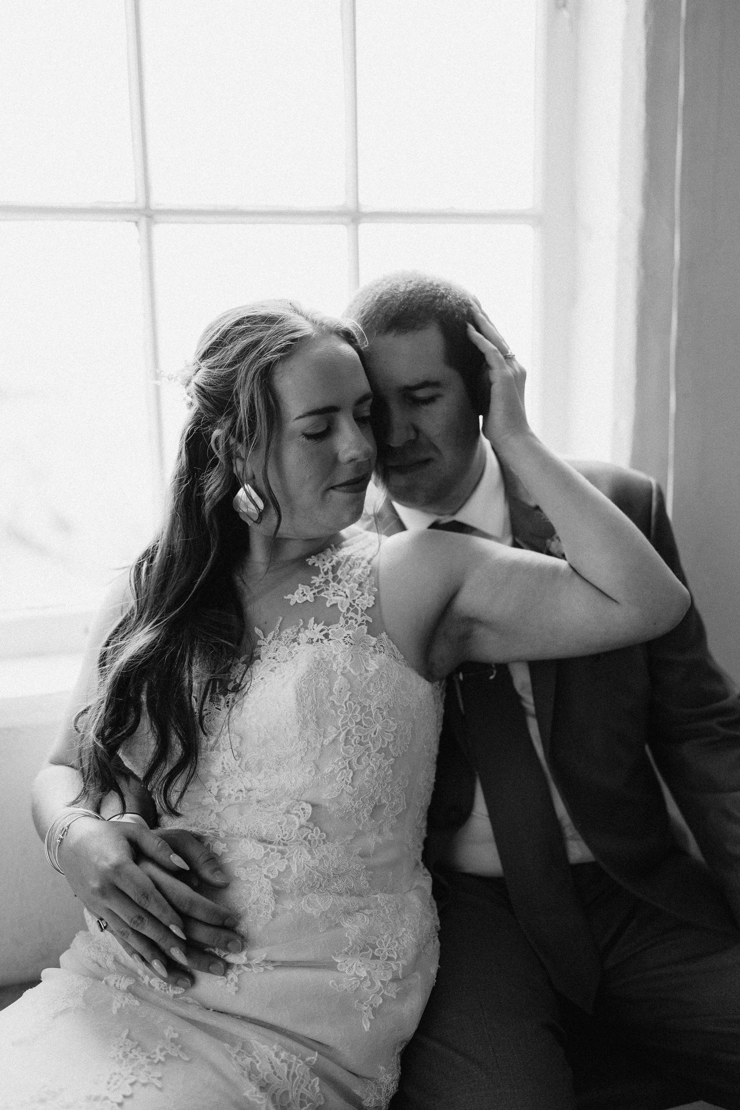 A romantic black and white portrait of a couple in wedding attire by a bright window.