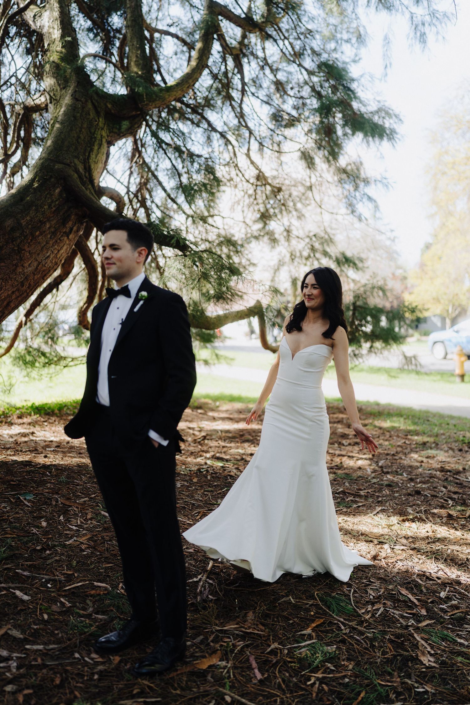 A bride in a white dress and groom in a black suit stand apart under a large tree in a park setting.