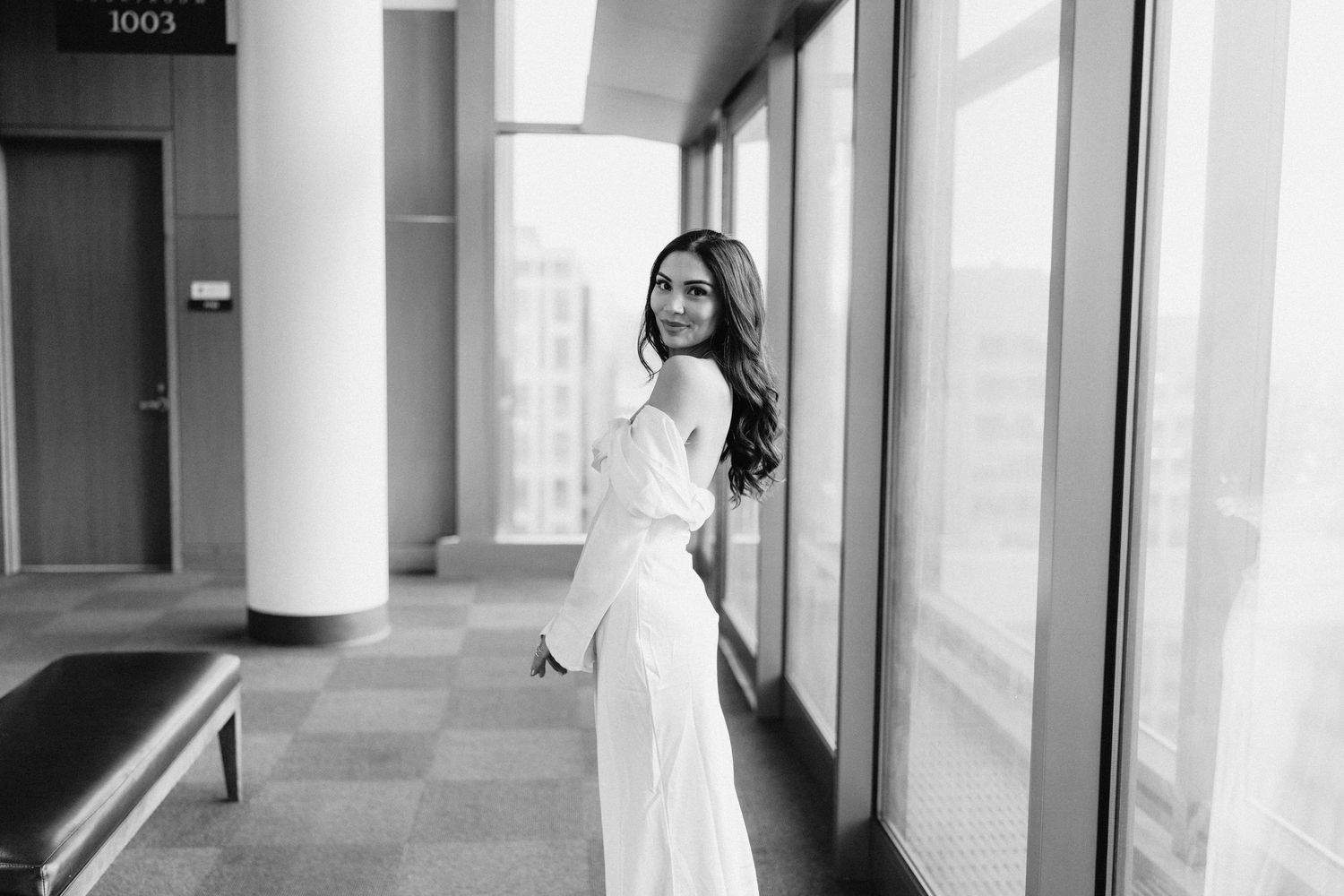 An elegant bride in an off-shoulder white dress poses by floor-to-ceiling windows in a modern building.