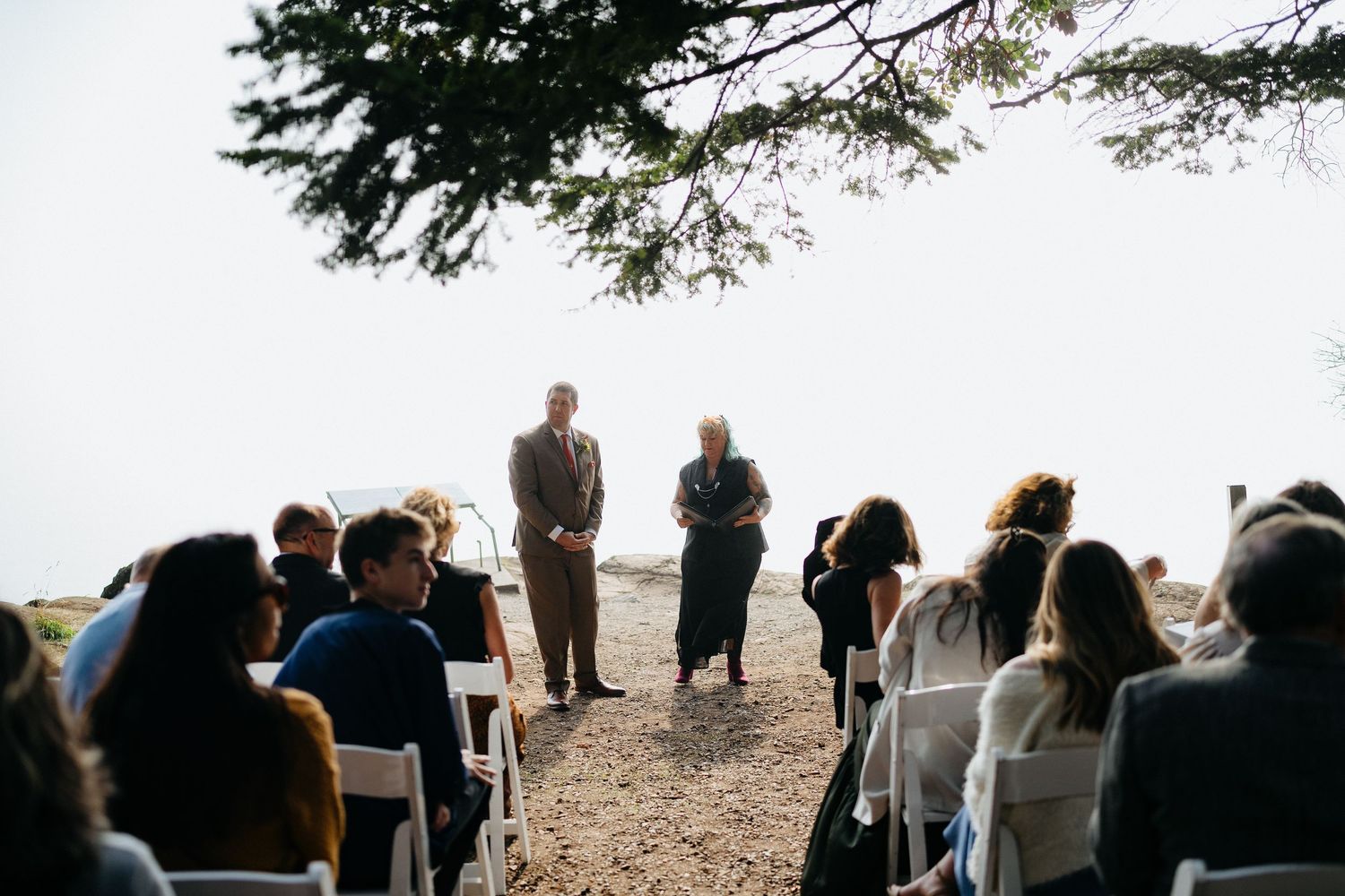 An outdoor wedding ceremony takes place on a cliff's edge overlooking a scenic natural landscape in late afternoon light.