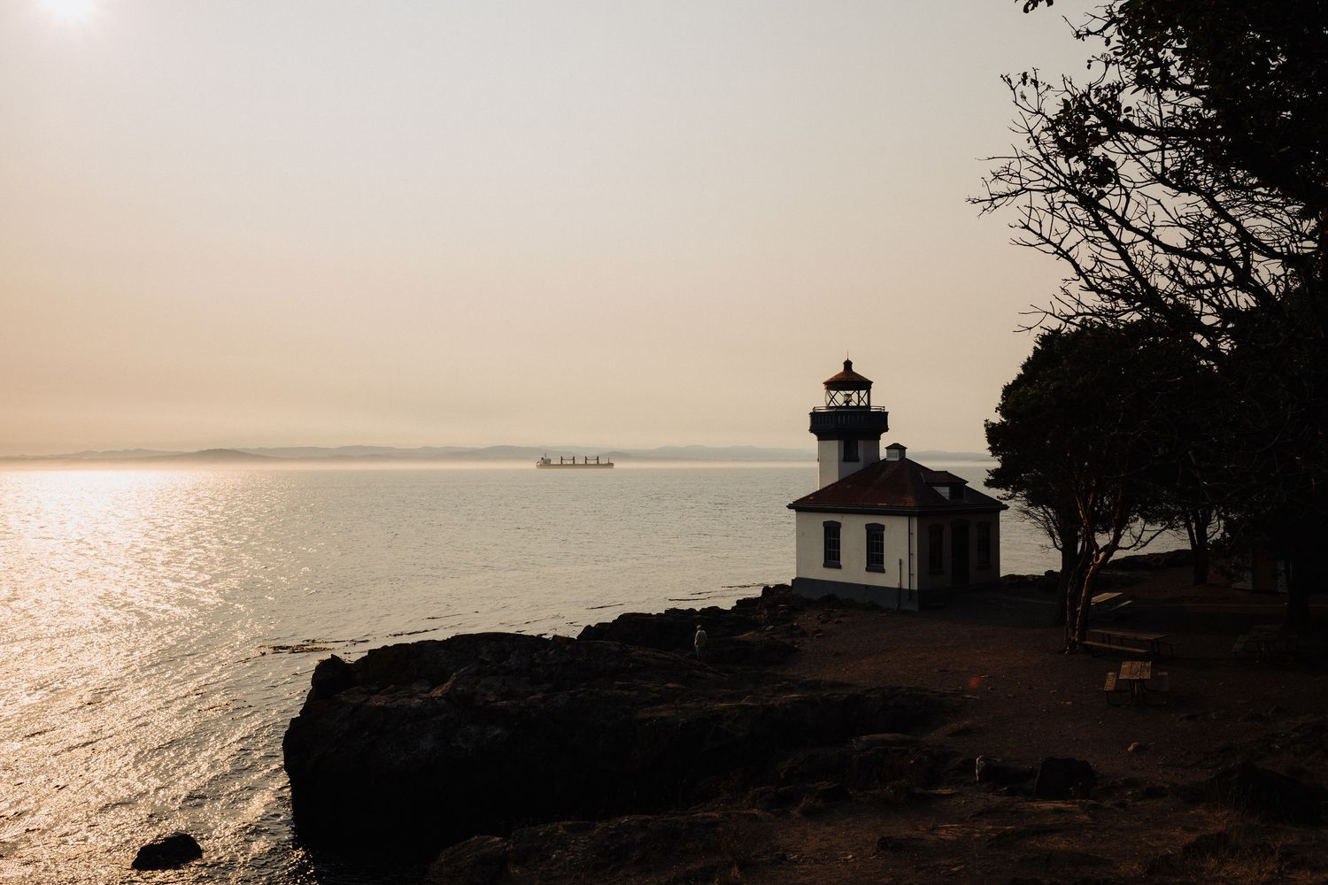 Lighthouse on rocky cliff overlooks serene ocean at sunset with glowing sunlight reflecting on calm waters.