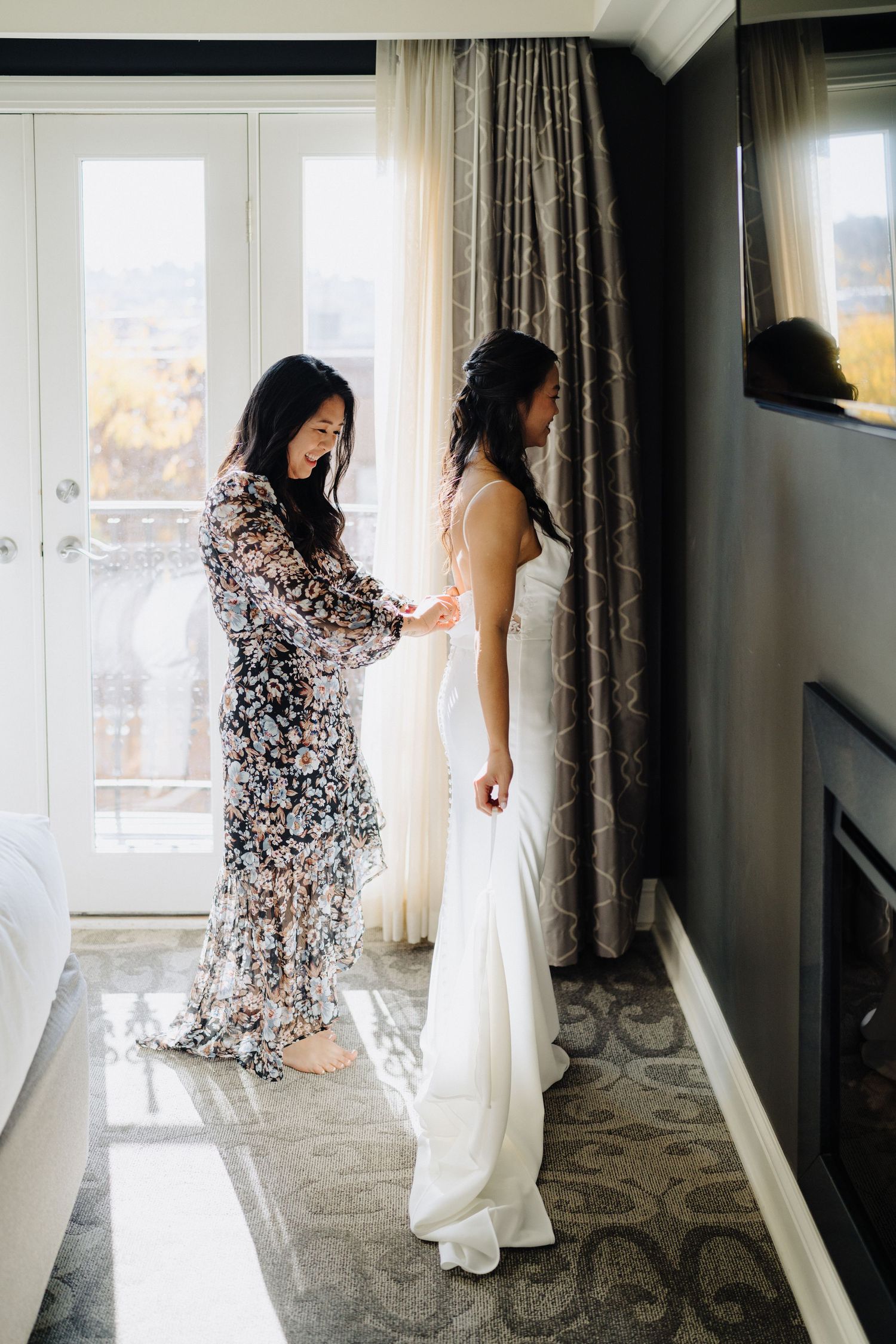 Two figures in a sunlit room preparing for a wedding, one in a floral dress helping adjust a white wedding gown.