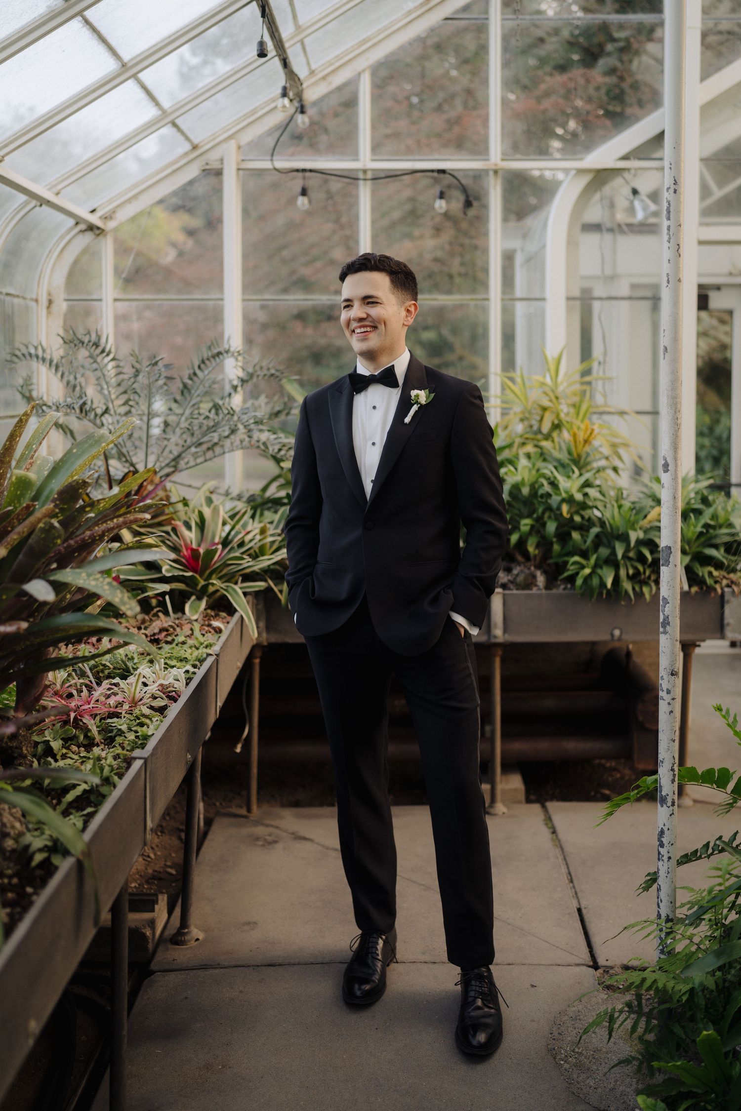 Groom in classic black tuxedo smiling in a greenhouse filled with tropical plants.
