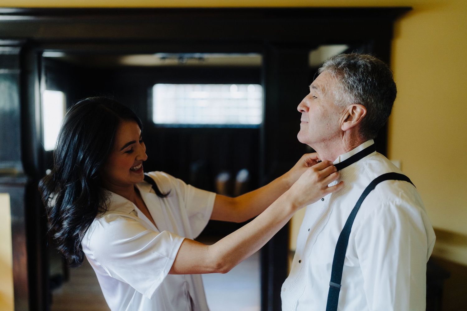 A person adjusts a bowtie and collar on someone wearing suspenders in an indoor setting.
