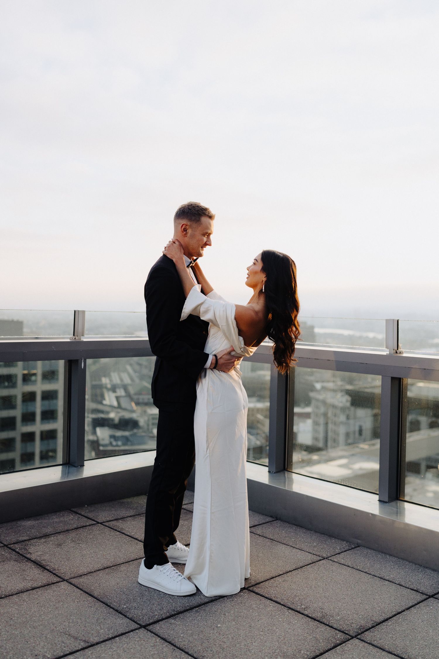 A romantic couple embraces on a high-rise balcony overlooking a cityscape at dusk.