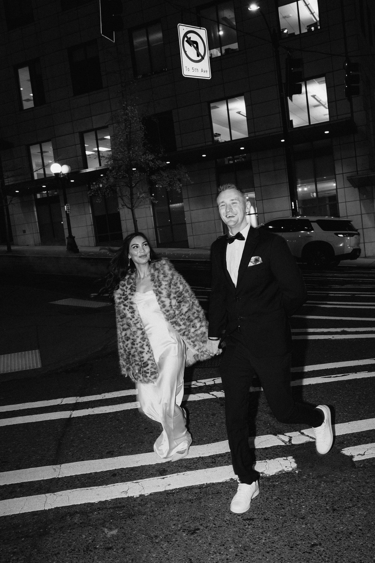 A stylish couple in formal wear crossing a city street at night.