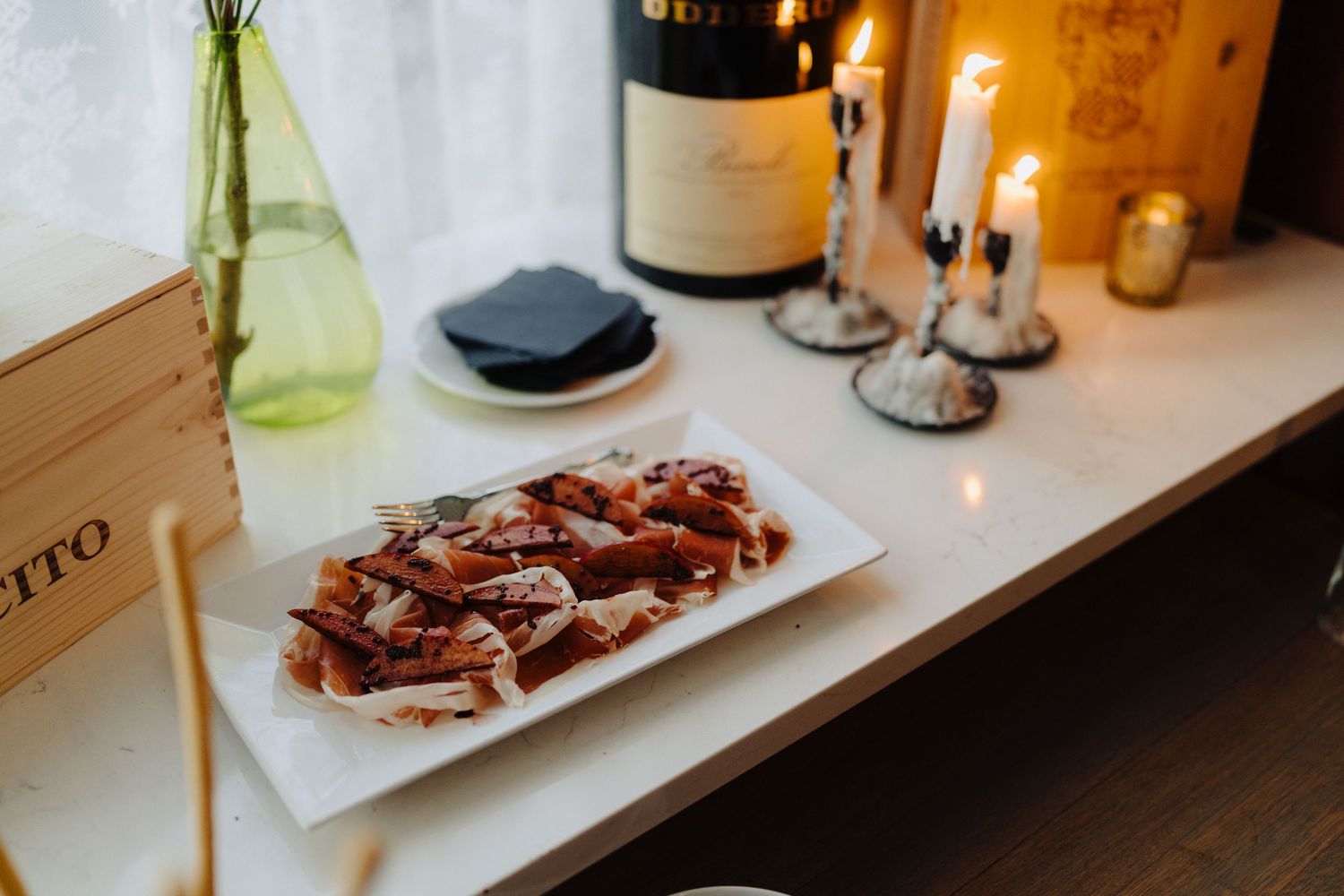 Plate of sliced meat appetizers alongside wine and candlesticks on wooden surface.