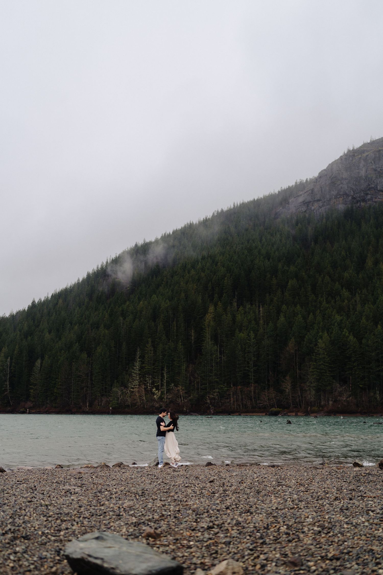 Two people stand together on a remote beach surrounded by fog-covered mountains and forest.