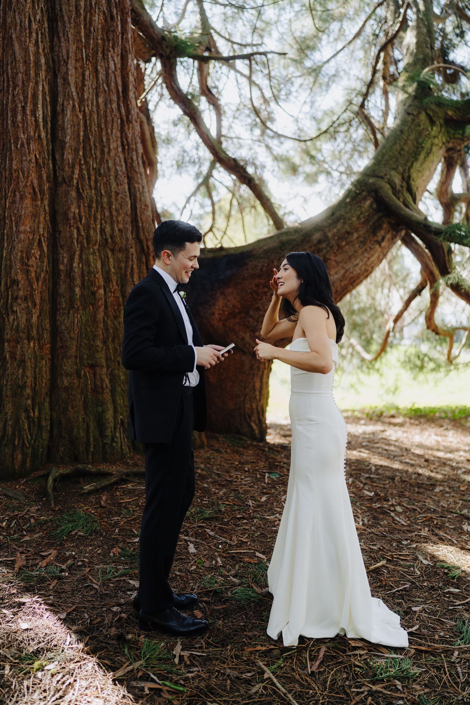 A couple shares an emotional first look moment beneath a towering redwood tree in a forest setting.
