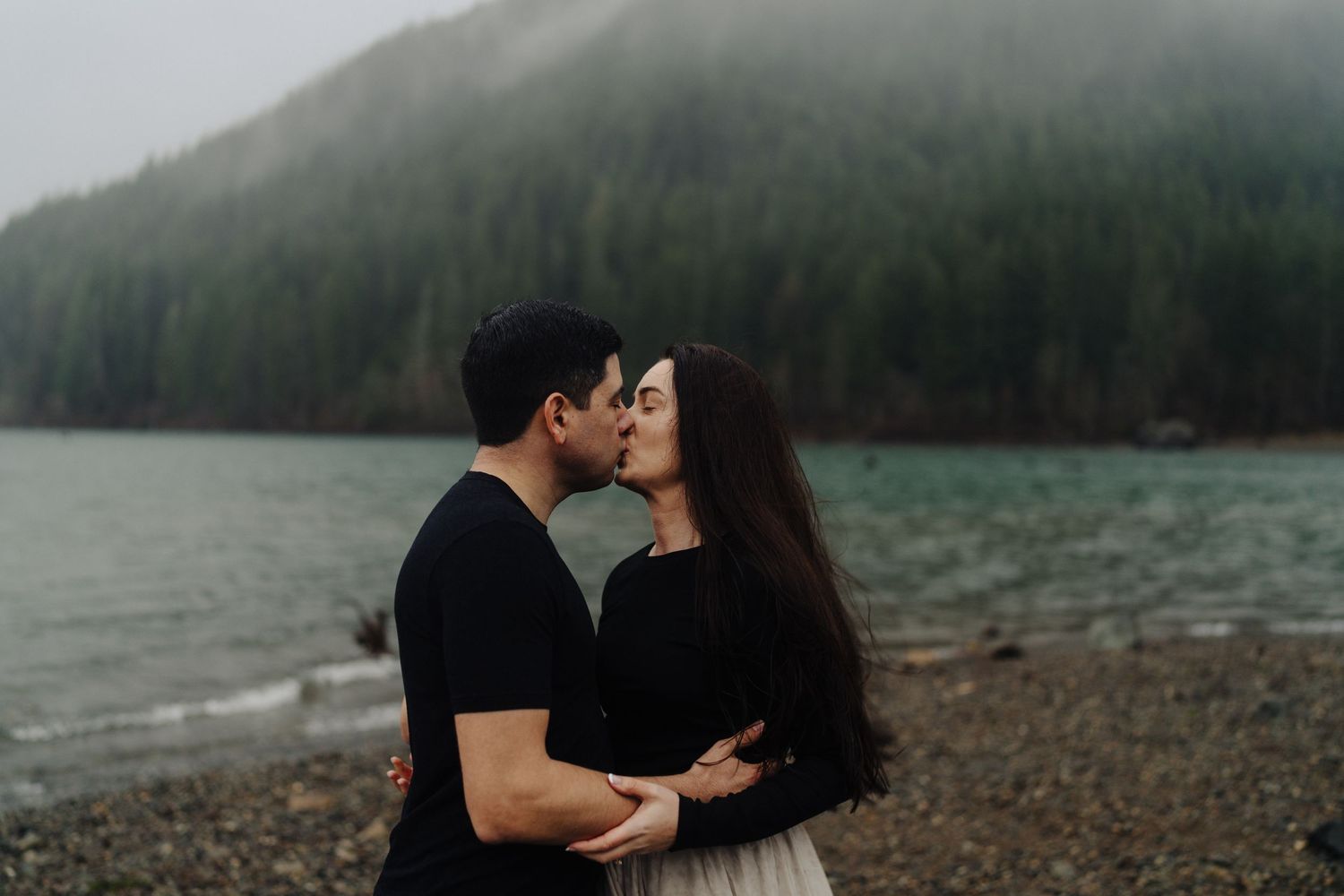 Two people in dark clothing share an intimate moment on a rocky beach with misty mountains and a lake in the background.