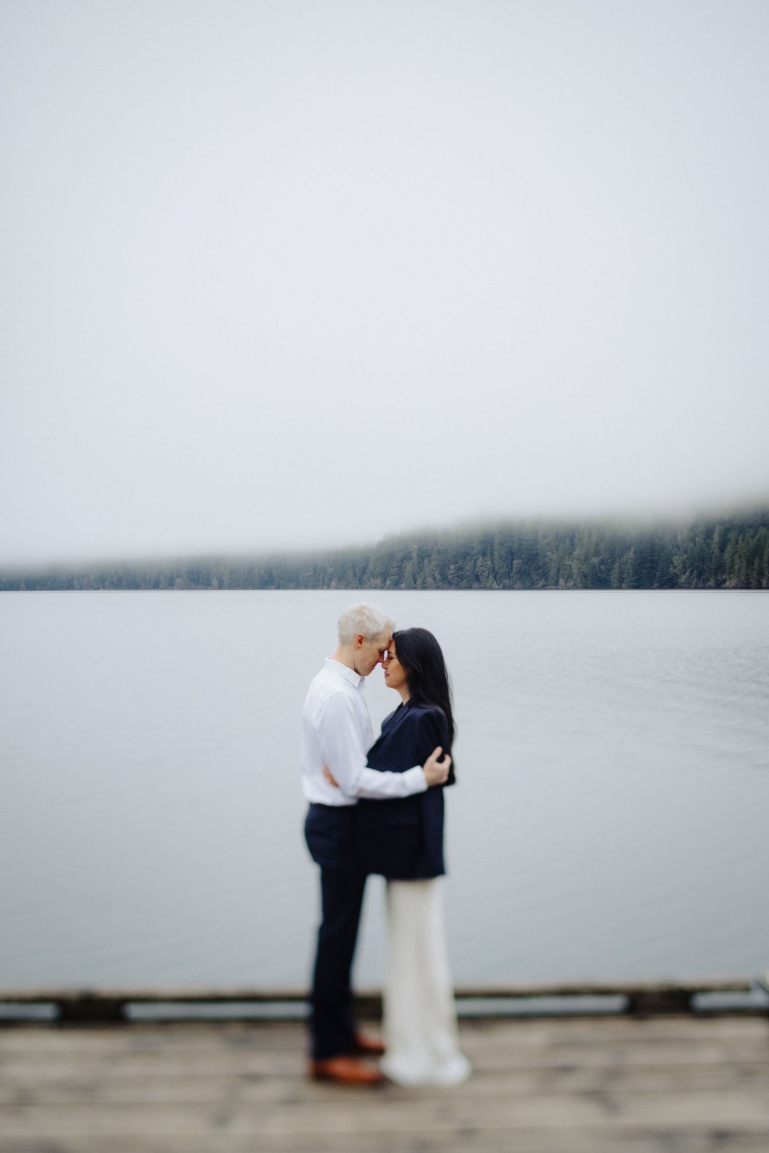 A couple shares a romantic moment on a misty lake dock with fog-covered mountains in the background.