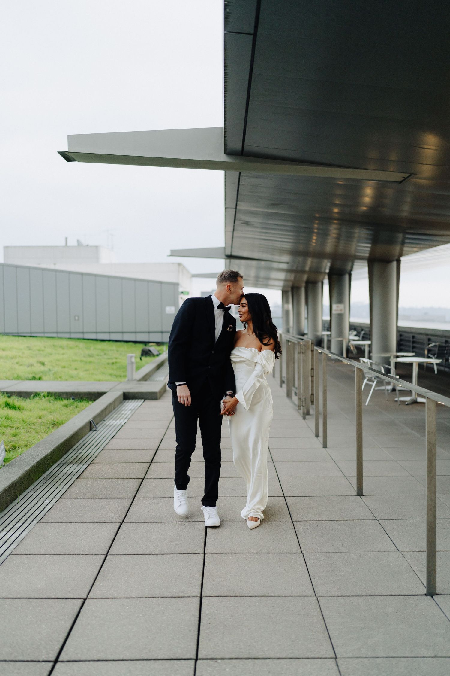A couple in wedding attire walking together under a modern architectural overhang.