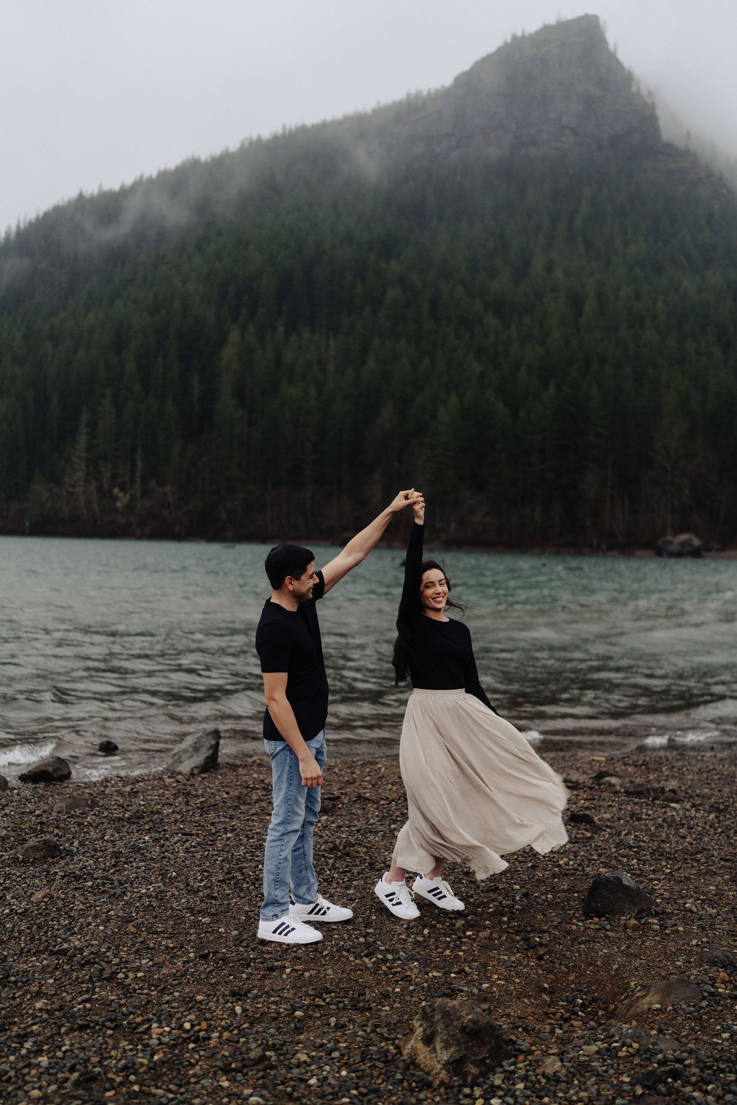 A couple dances on a rocky beach with fog-covered mountains and evergreen forest in the background.