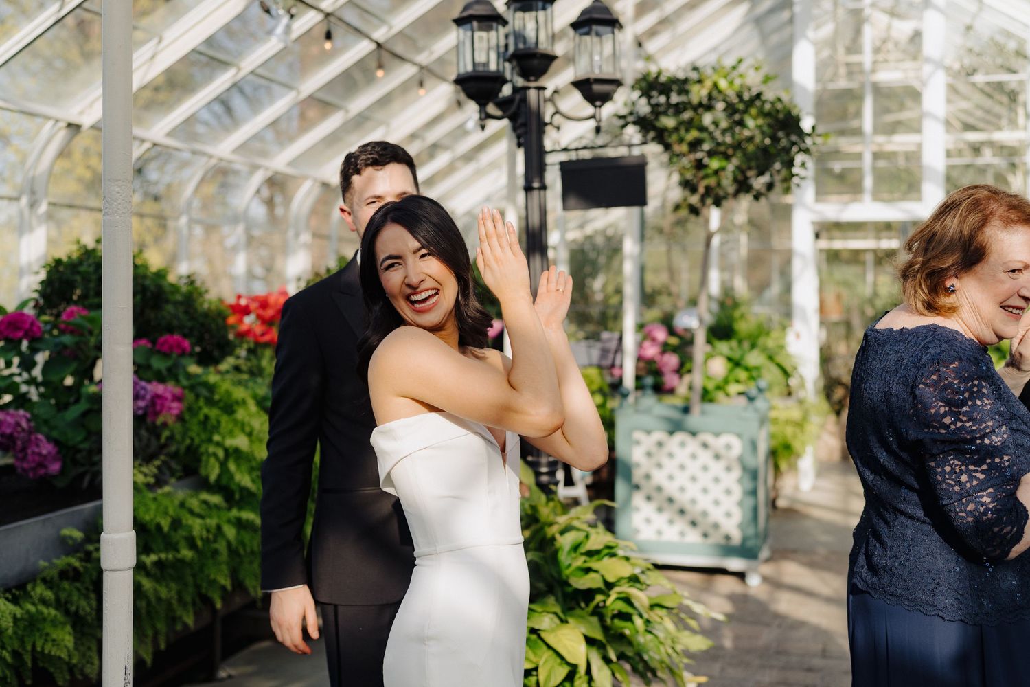 A joyful celebration moment captured in a greenhouse wedding venue with hanging lights.