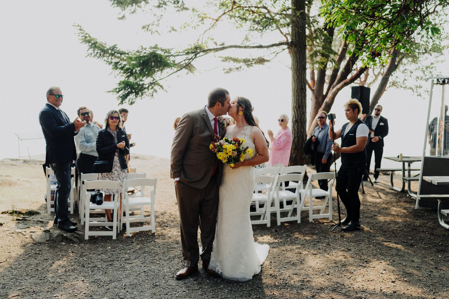 A couple embraces during an intimate outdoor wedding ceremony with guests and white chairs arranged under trees by the water.