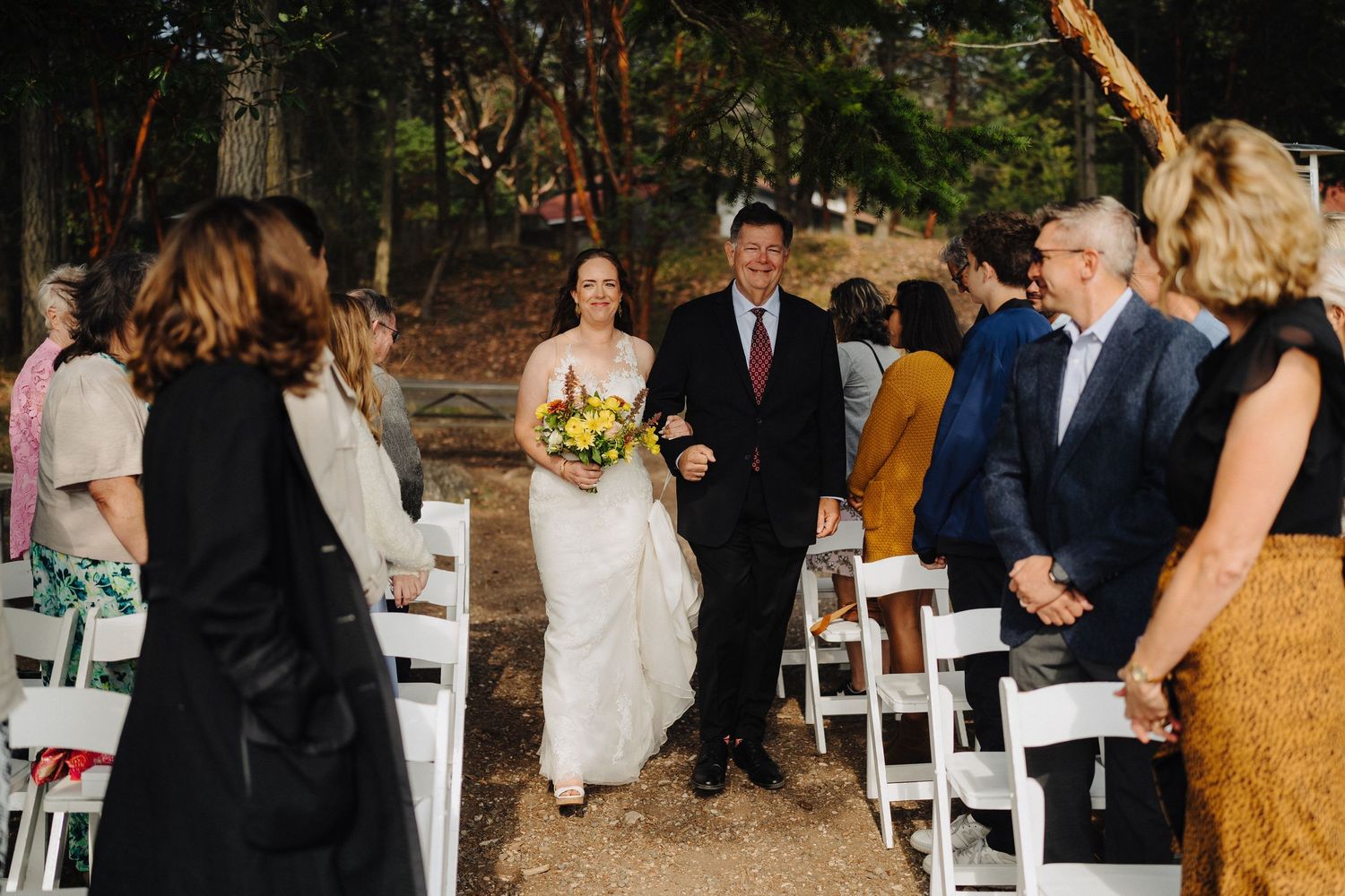 A wedding ceremony takes place outdoors with guests seated in white chairs and greenery in the background.