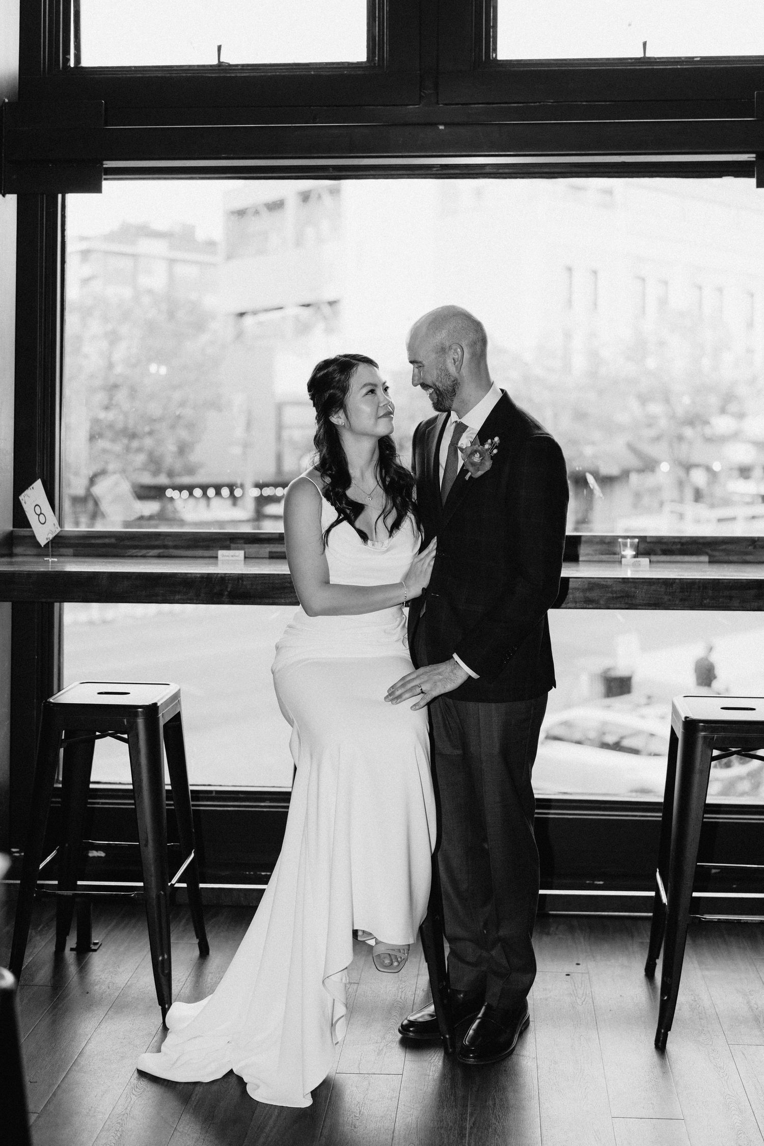Black and white portrait of a couple in wedding attire standing by large windows overlooking a city view.