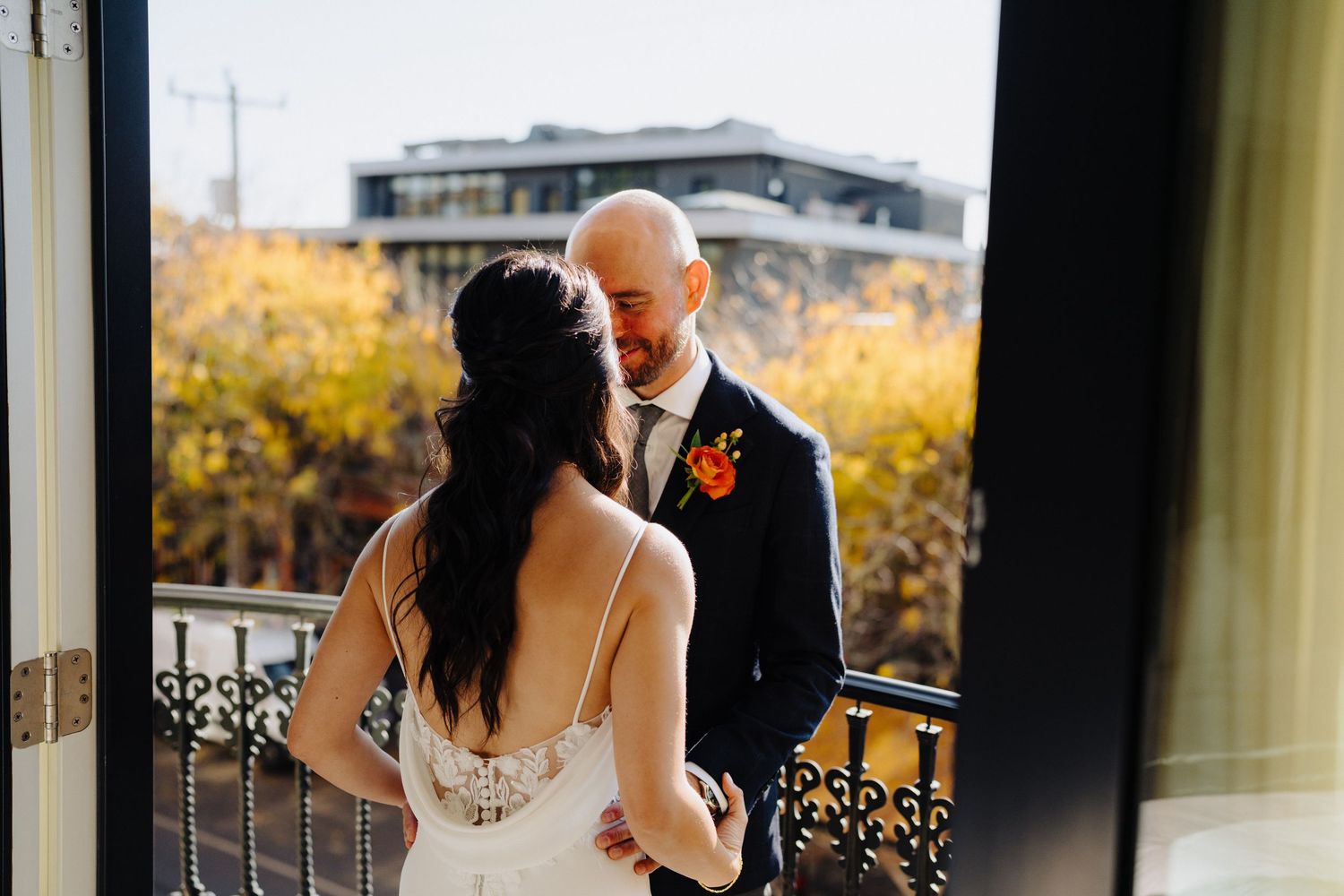 A romantic wedding couple shares an intimate moment on a balcony with autumn foliage in the background.
