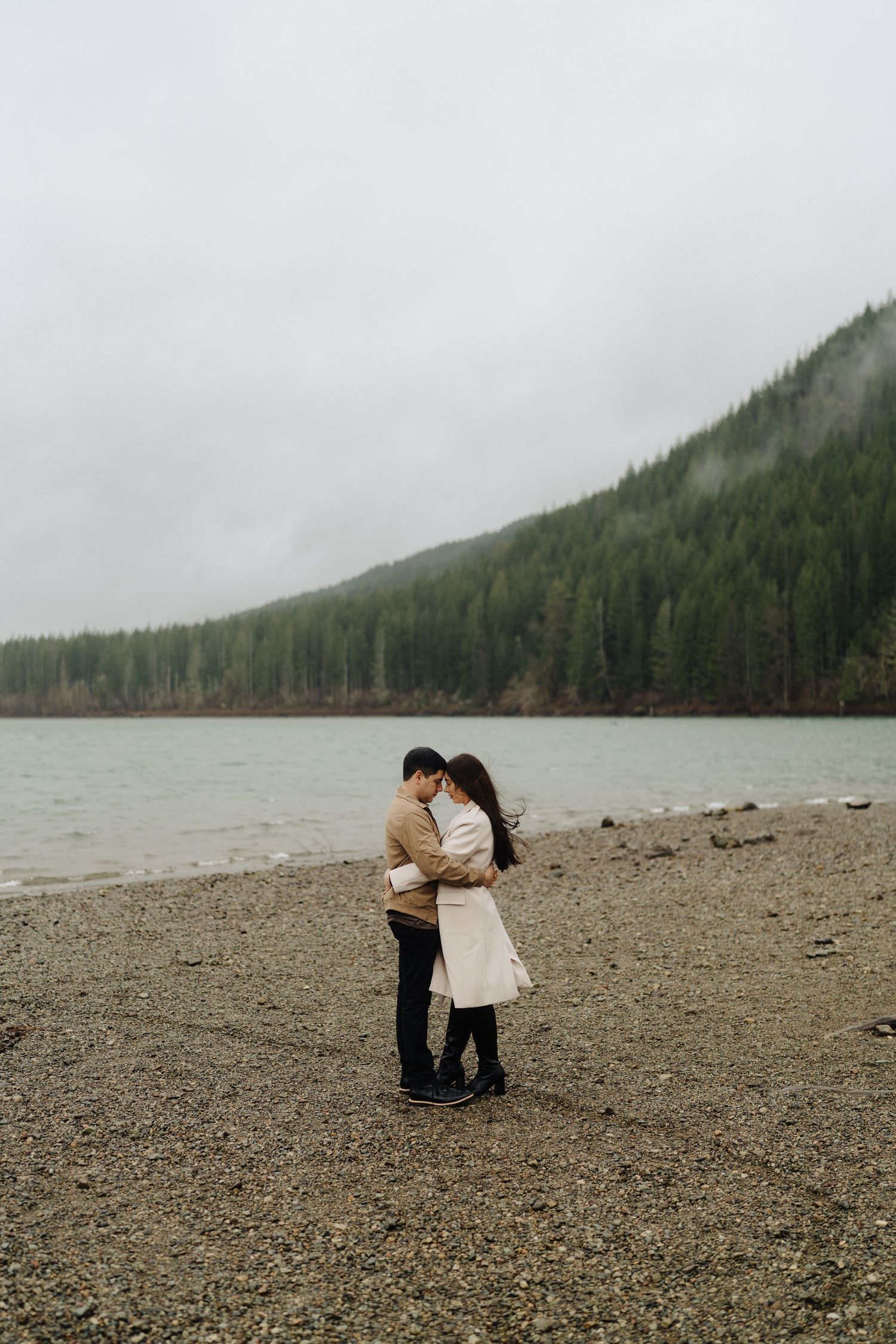 A couple embraces on a rocky beach shore with misty mountains and evergreen forest in the background.