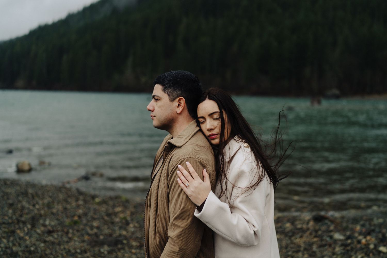 A couple shares an intimate moment by a moody mountain lake with evergreen forests and fog.