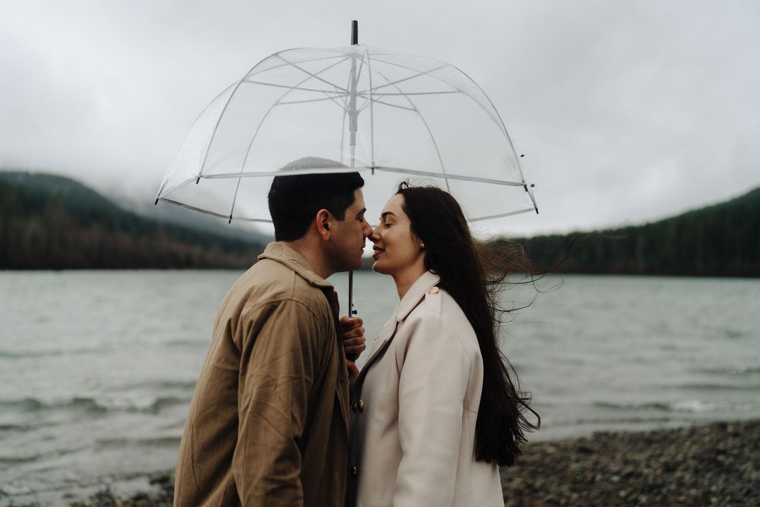 A romantic couple shares an intimate moment under a clear umbrella by a misty lake on an overcast day.