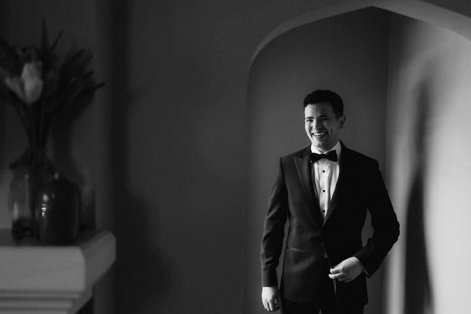 An elegant groom fastens his bowtie while getting ready in a classic black and white photograph.