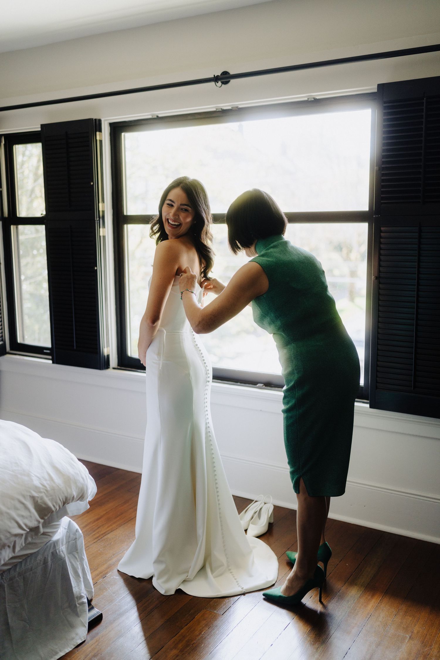 A tender moment of getting dressed in a sleek white wedding gown by a window with dark shutters.