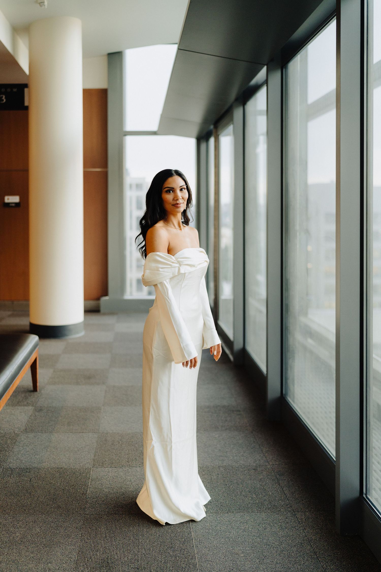 Elegant white off-shoulder gown photographed in modern building hallway with floor-to-ceiling windows.