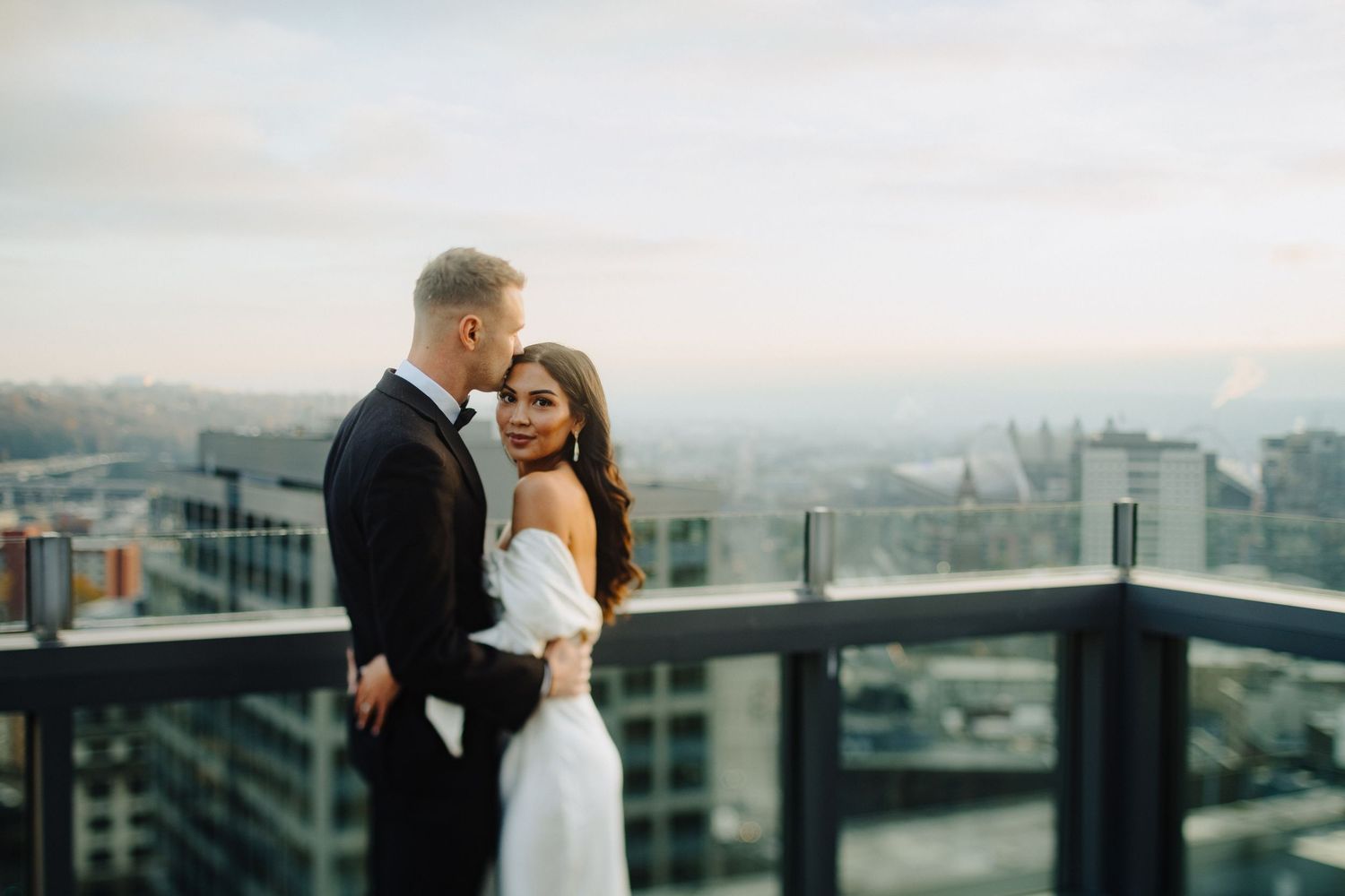 A couple embraces on a rooftop terrace with a cityscape backdrop during sunset.