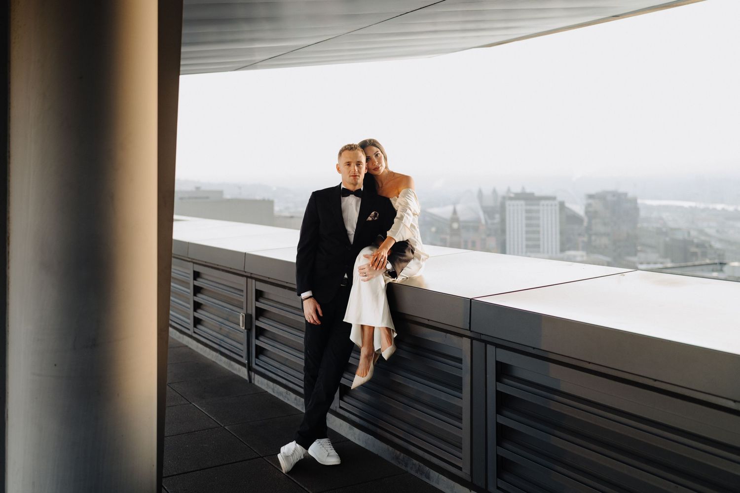A couple in formal attire shares a romantic moment on a high-rise balcony overlooking the city.