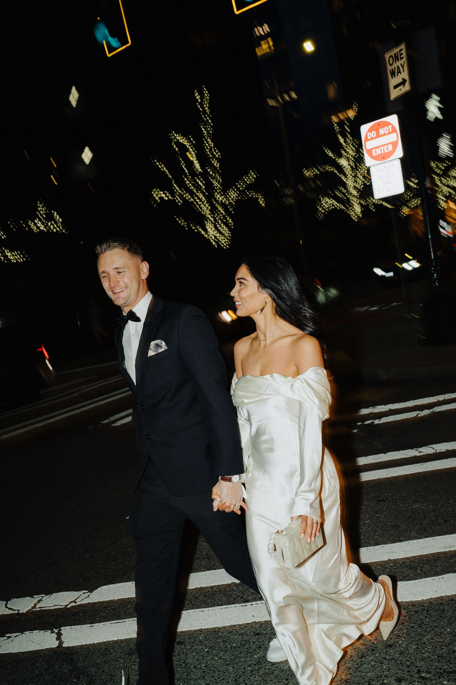 A couple in wedding attire walks hand in hand across a crosswalk at night with city lights glowing behind them.