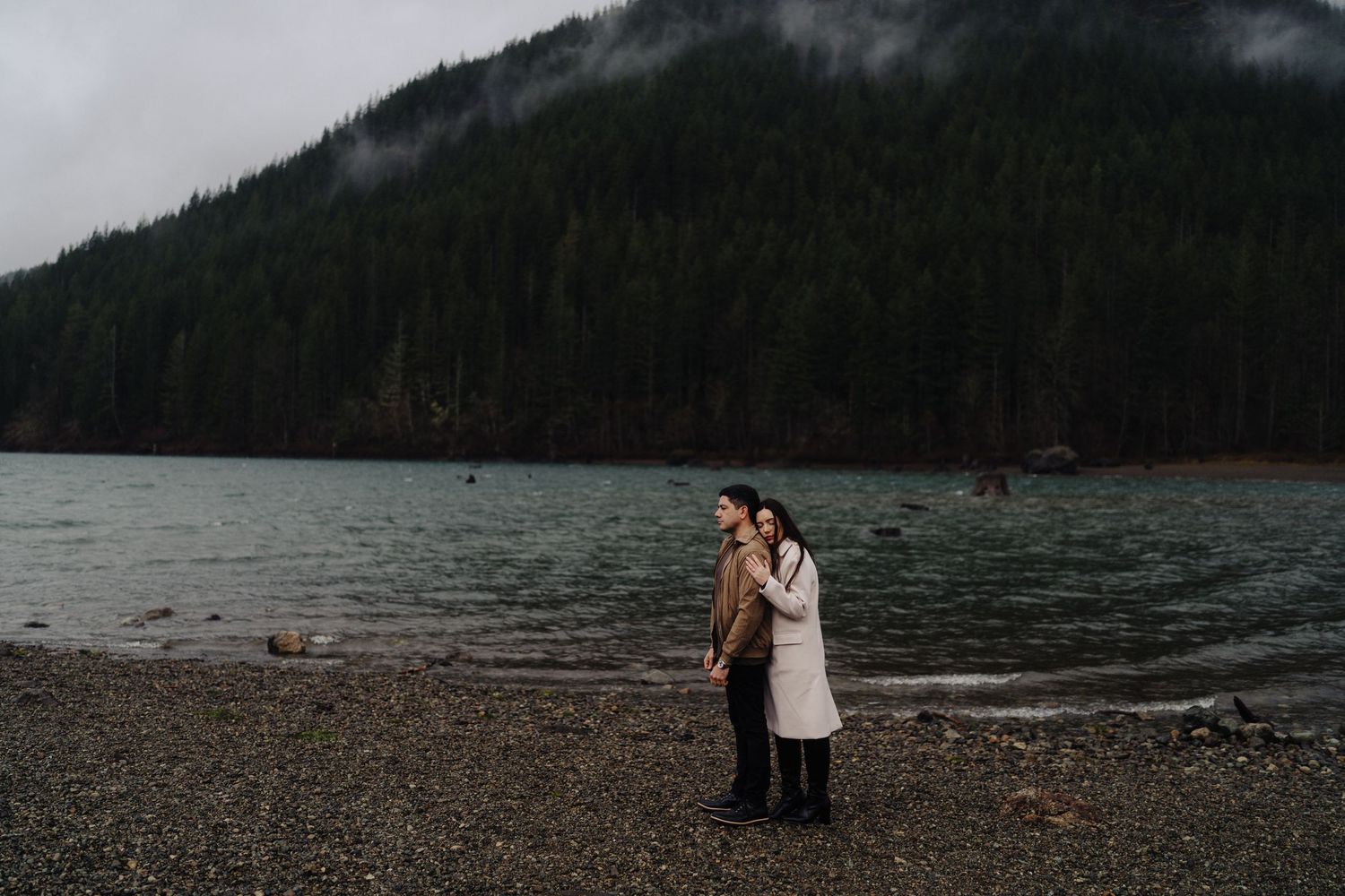 Couple stands together on rocky beach with evergreen forest and mountains creating a moody backdrop.