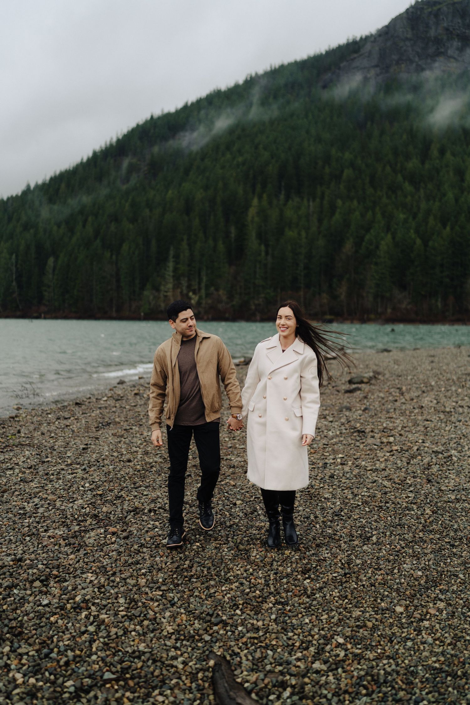 A couple walks together on a pebble beach against a backdrop of foggy mountains and evergreen forest.