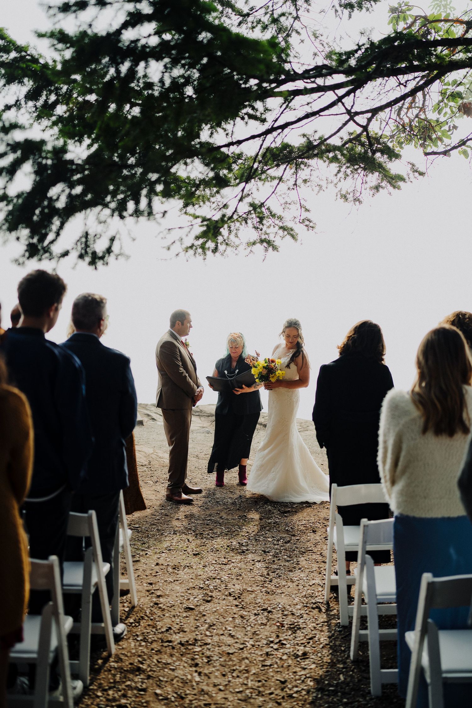 An intimate outdoor wedding ceremony overlooking the ocean with guests seated on white chairs beneath overhanging trees.