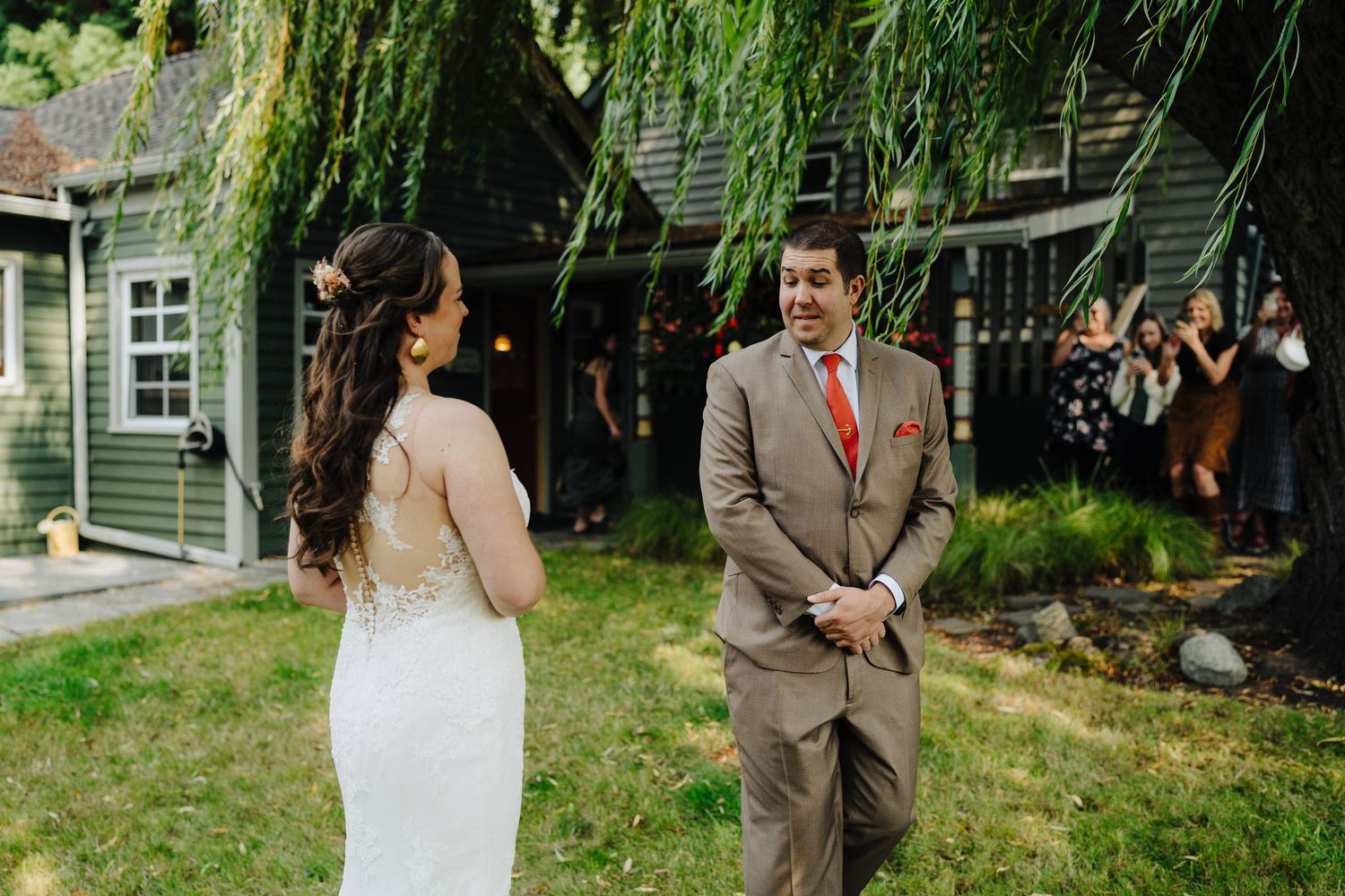 A couple in wedding attire stands together in a garden with a willow tree and green house in the background.
