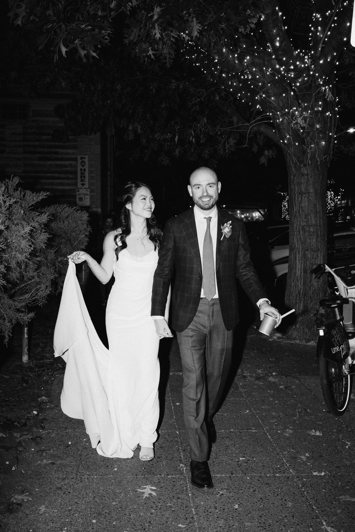 A couple walking under twinkling lights on a nighttime street in formal attire.