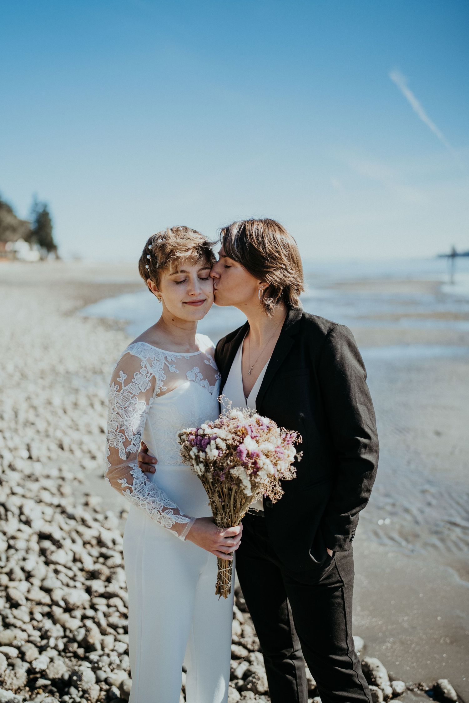 A romantic beach wedding couple shares a tender moment on a pebbled shore holding a bouquet of pink flowers.