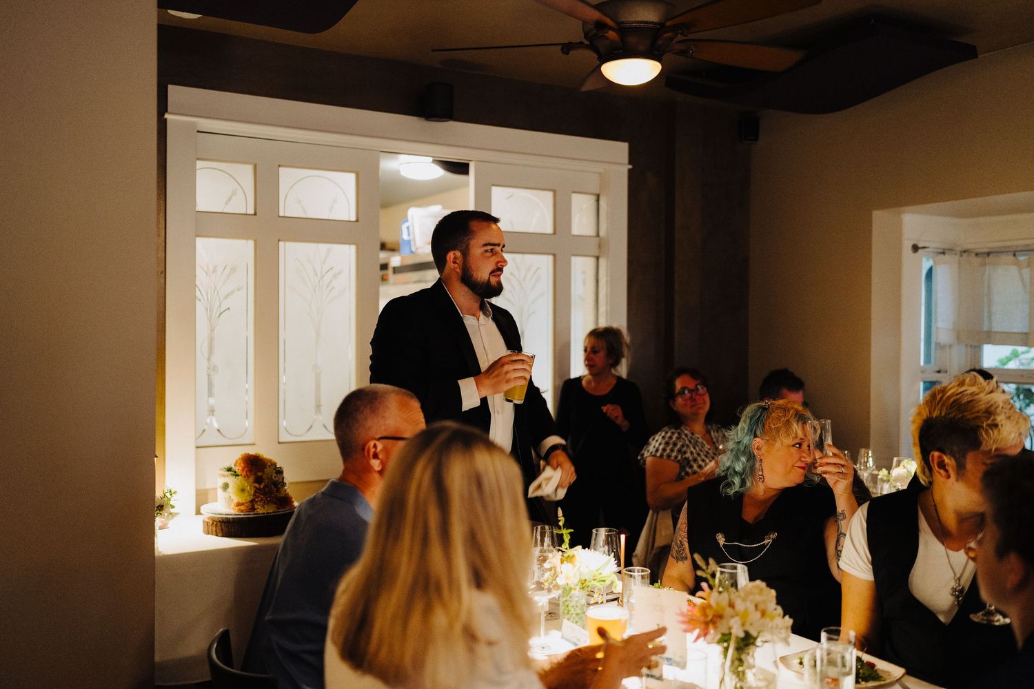 Wedding guests enjoy a reception dinner in an elegant indoor venue with large windows.