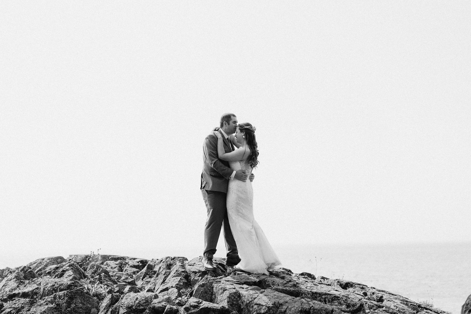 A romantic black and white photo shows a couple embracing while standing on a rocky mountaintop overlook.