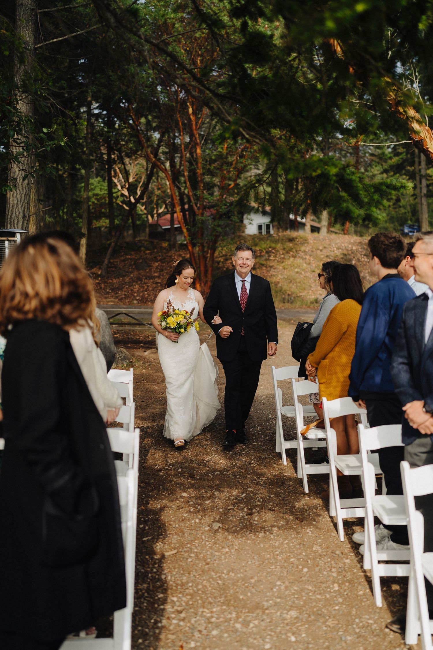 Wedding guests seated on white chairs in a wooded outdoor ceremony setting.