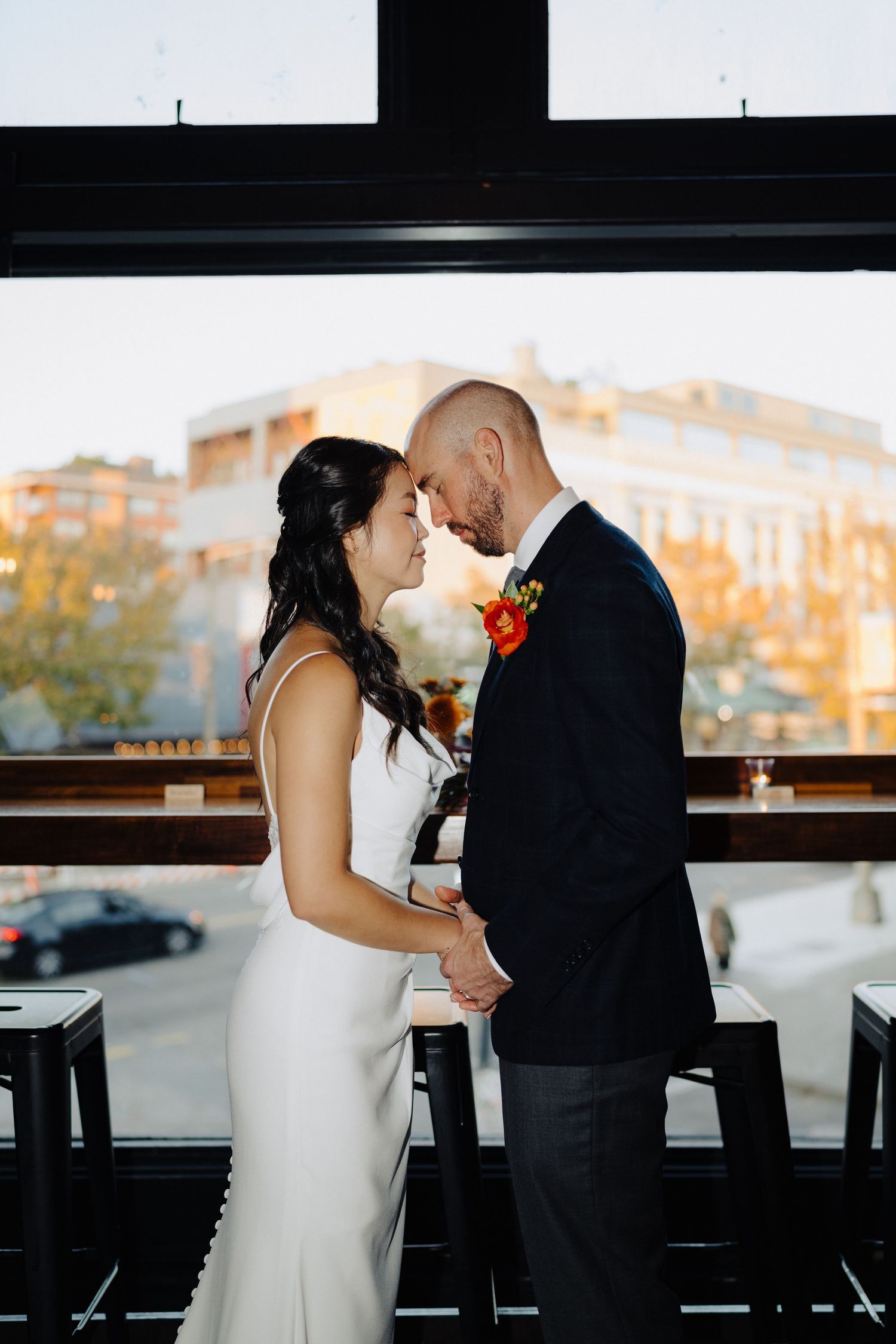 Romantic moment between newlyweds by window during sunset at urban venue.