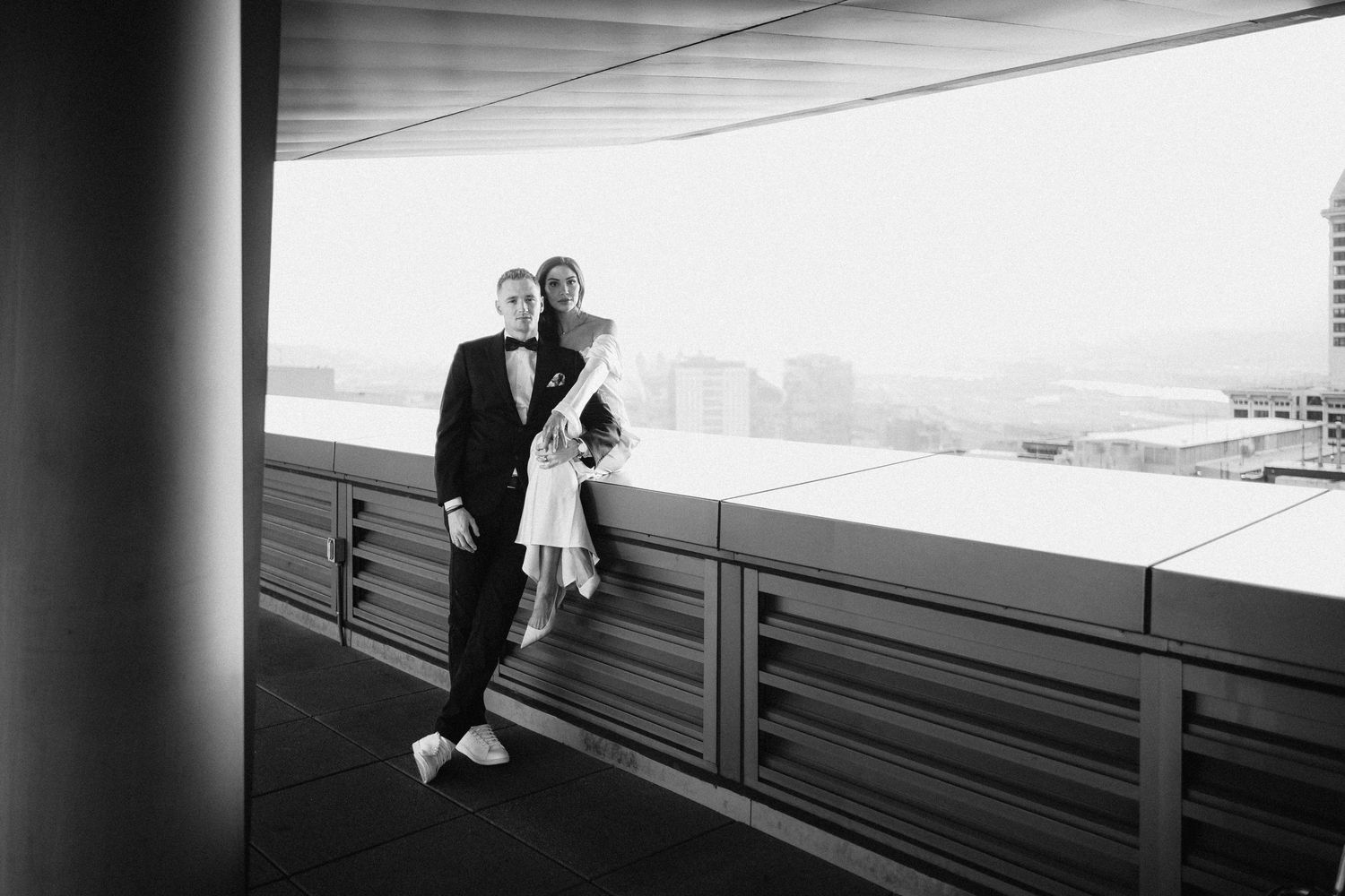 A black and white photo of a couple standing on a modern balcony overlooking the city.