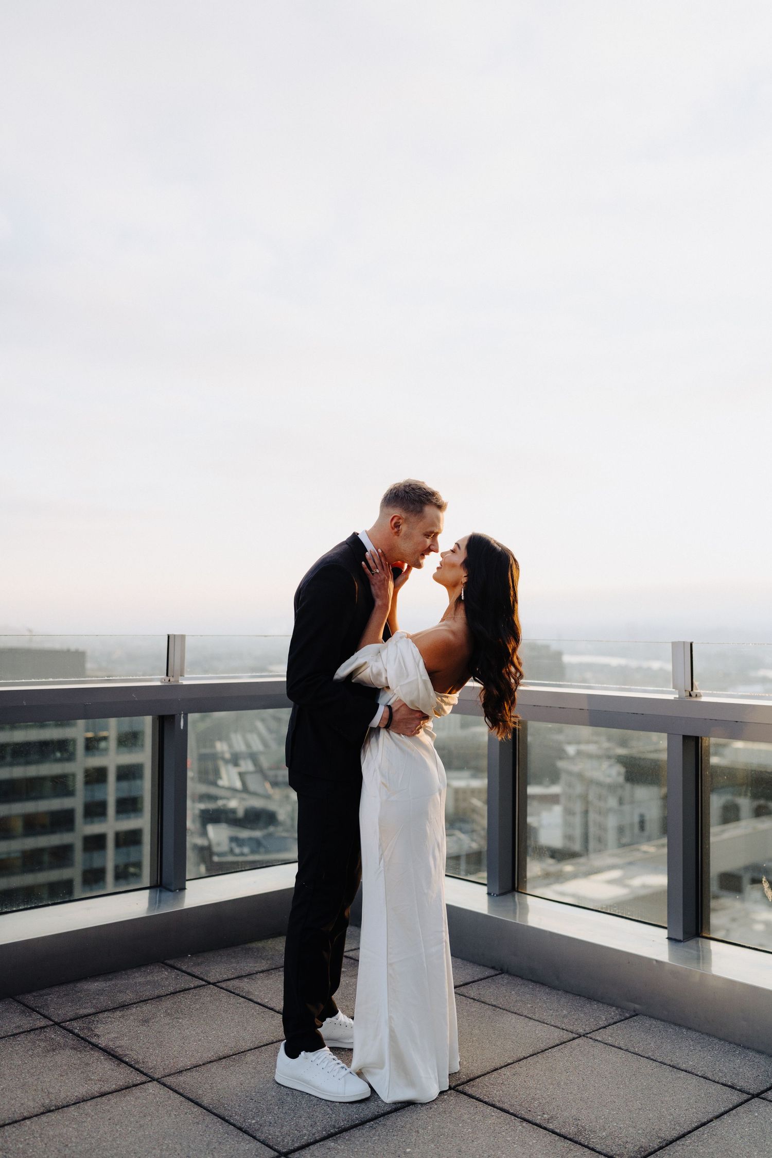 A couple shares a romantic moment on a high-rise balcony overlooking the city, dressed in formal wedding attire.