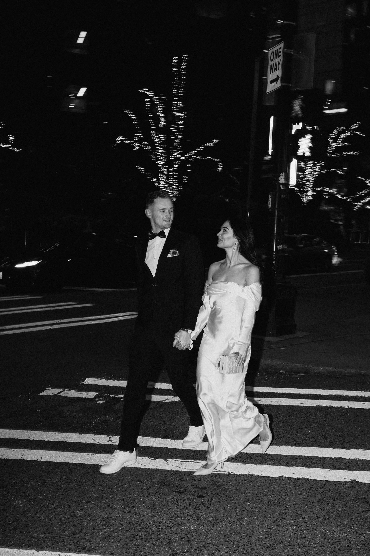 A black and white photo of a couple walking across an illuminated city crosswalk at night.