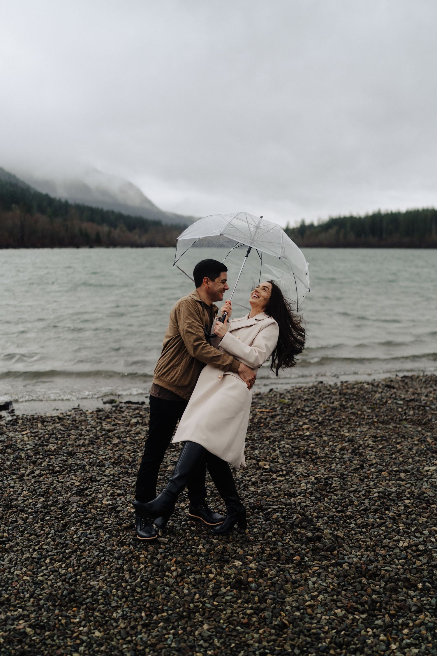 Couple sharing umbrella moment on rainy beach with misty mountain landscape.