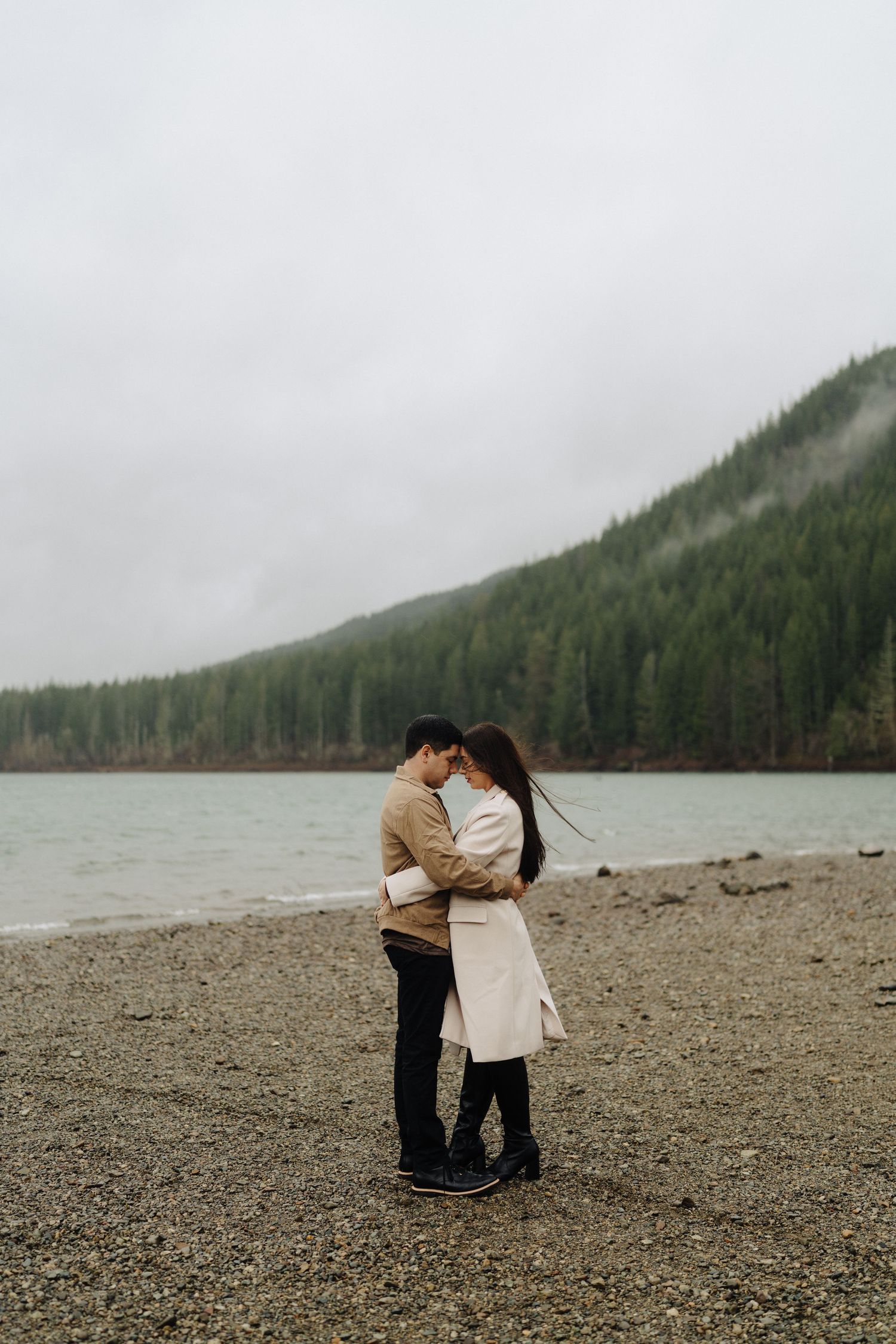 A couple embraces on a rocky beach shore with misty mountains and evergreen forest in the background.