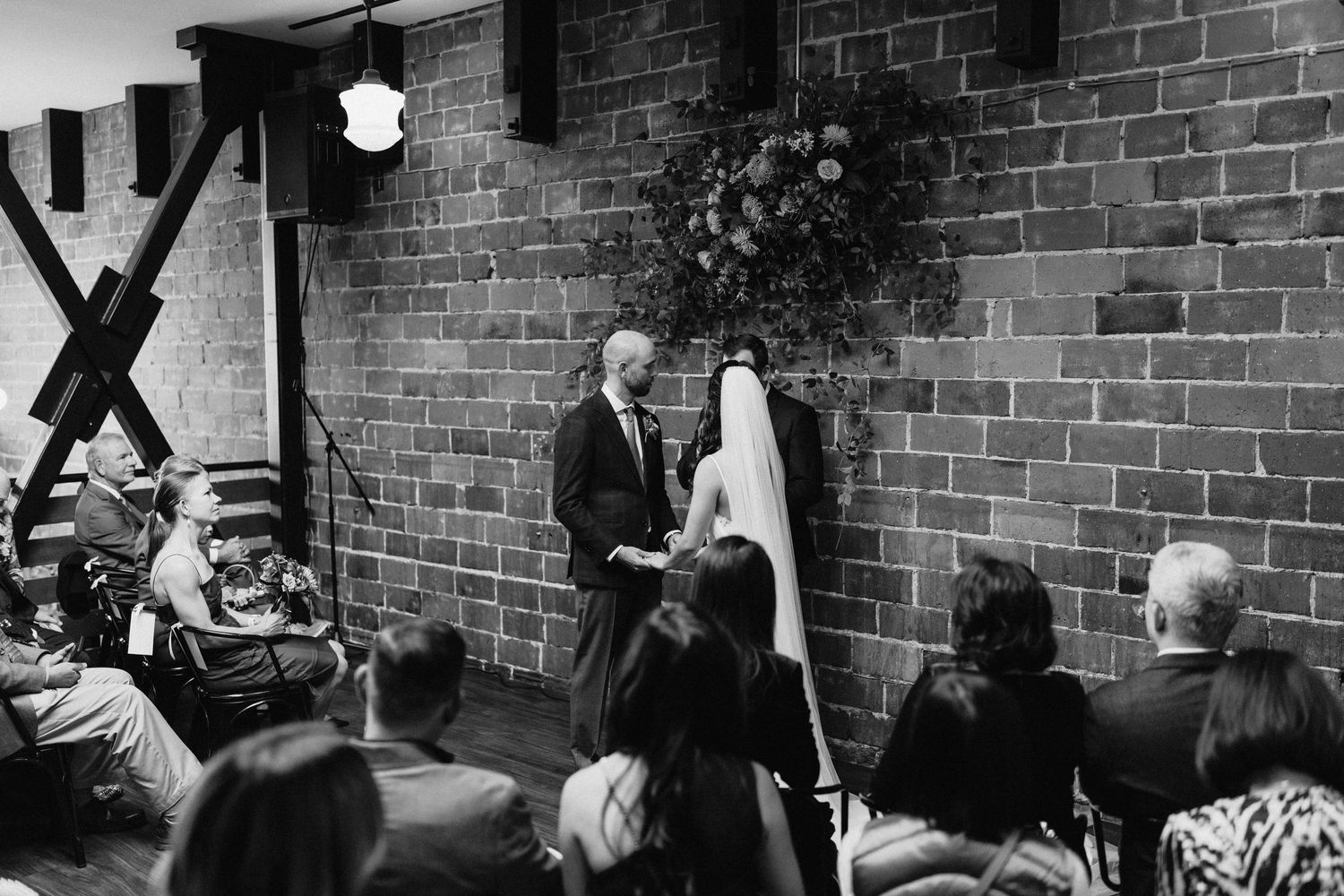 A black and white wedding ceremony captured in front of an industrial brick wall setting.