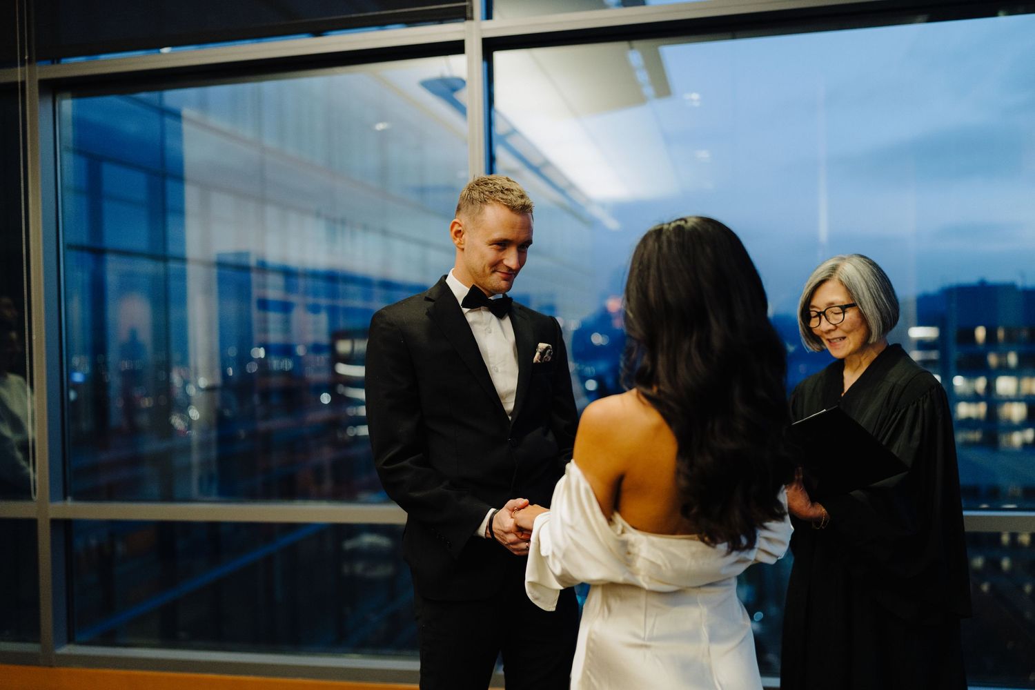 An intimate wedding ceremony takes place at dusk in a modern high-rise venue with city views in the background.